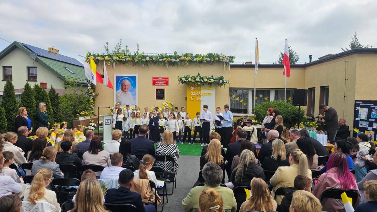 Na zdjęciu Jubileusz 20-lecia nadania imienia Jana Pawła II Przedszkolu nr 9. Uczestnicy uroczystości siedzą na placu przedszkolnym, wkoło barwy papieskie i chorągiewki