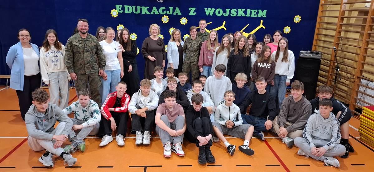 Na zdjęciu uczestnicy programu Edukacja z wojskiem w Szkole Podstawowej nr 13. Pozują do wspólnego zdjęcia z wojskowymi prowadzącymi zajęcia. W tle hasło Edukacja z wojskiem