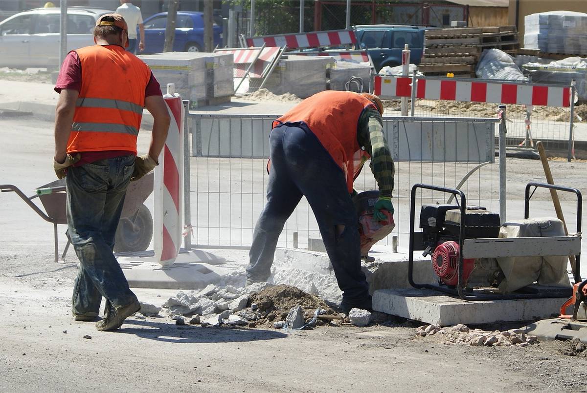 Na zdjęciu robotnicy drogowi w pomarańczowych kamizelkach remontują drogę przy użyciu sprzętu mechanicznego