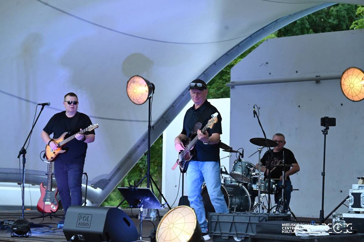 Na zdjęciu trio rockowe Kazzu podczas występu w ramach Letniej Sceny Artystycznej w amfiteatrze Przystani nad Pilicą. Z przody dwóch gitarzystów z tyłu perkusista, wszyscy w czarnych t-shirtach