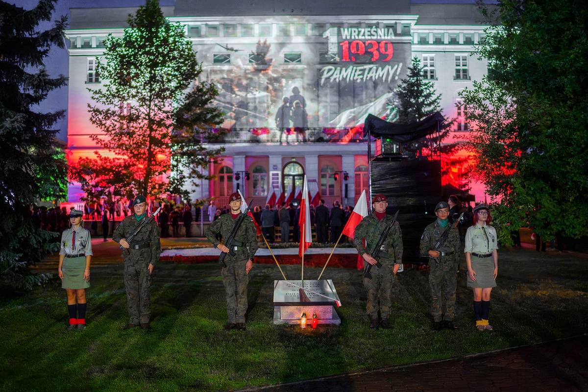 Warta honorowa przy płycie poświęconej pamięci Nieznanego Żołnierza. Na pierwszym planie umundurowani zołnierze oraz harcerze. Za nimi biało-czerwone flagi Polski. W tle fasada Urzędu Miasta z  okolicznościową projekcją: motyw płomieni, żołnierze itp.
