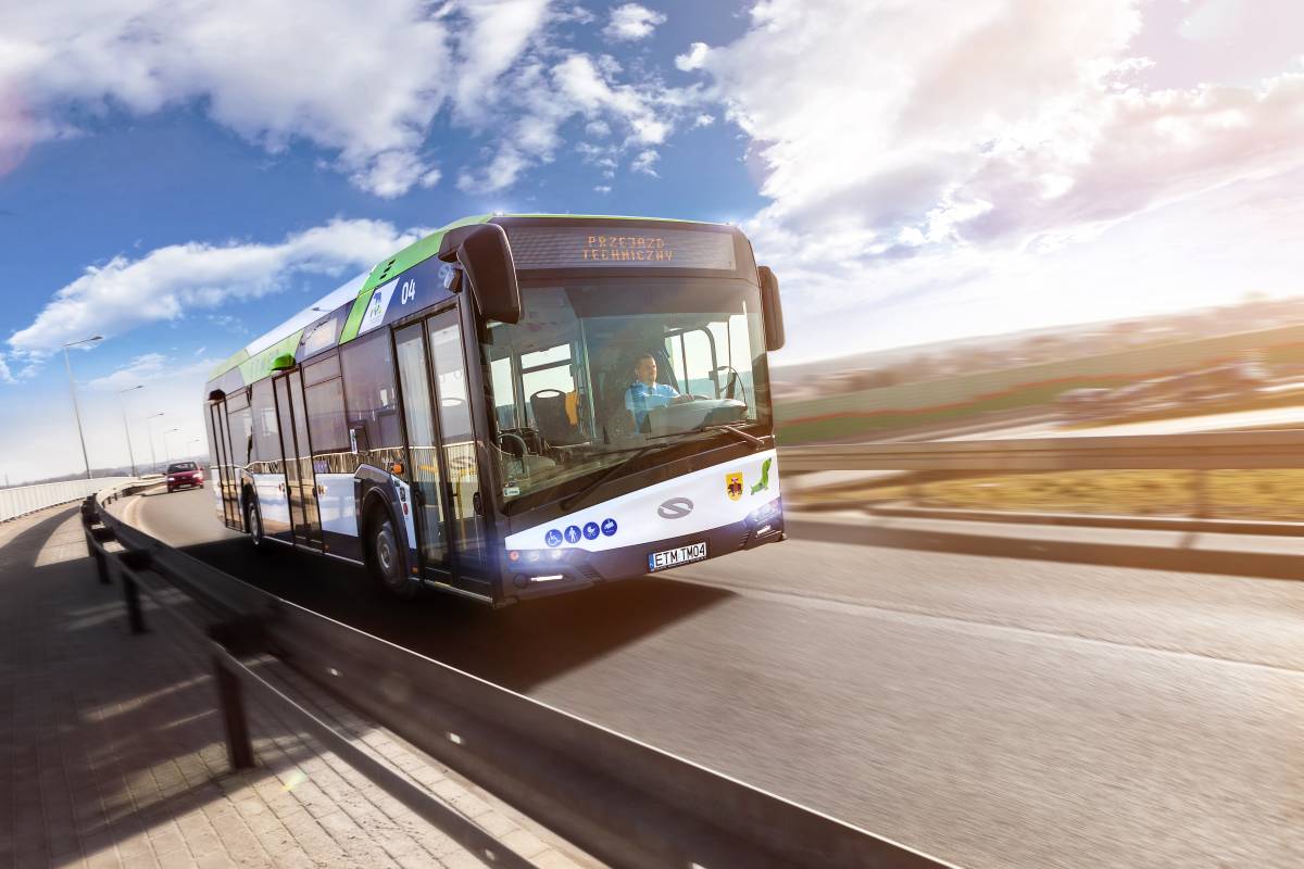 Niskopodłogowy autobus miejski, poruszający się na drodze asfaltowej. Jezdnię oddziela od ciągu pieszego metalowa bariera. Niebo jasnoniebieskie z rozproszonymi, białym chmurami.