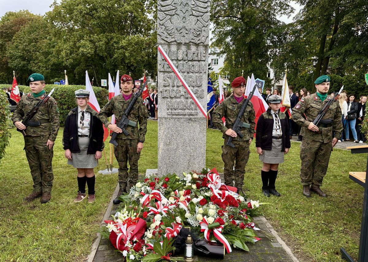 W centrum Pomnik „W Hołdzie Zesłańcom Sybiru”. Przy pomniku warta honorowa harcerzy i wojska. Obelisk zdobi biało-czerwona wstęga i złożone prze delegacje kwiaty. W tle uczestnicy z flagami narodowymi na drzewcach.