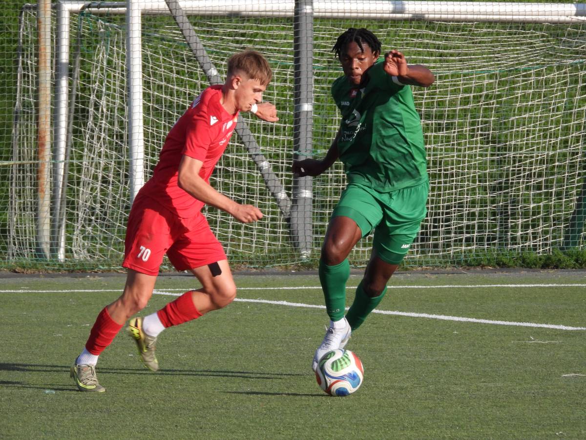 Mecz Lechia -Świt rozgrywany w słoneczny dzień na boisku z zieloną, sztuczną murawą. W tle widoczna metalowa konstrukcja bramki oraz siatka. Przy piłce zawodnik w zielonym stroju próbuje ominąć przeciwnika. Piłkarz Lechii w czerwonym stroju walczy o piłkę
