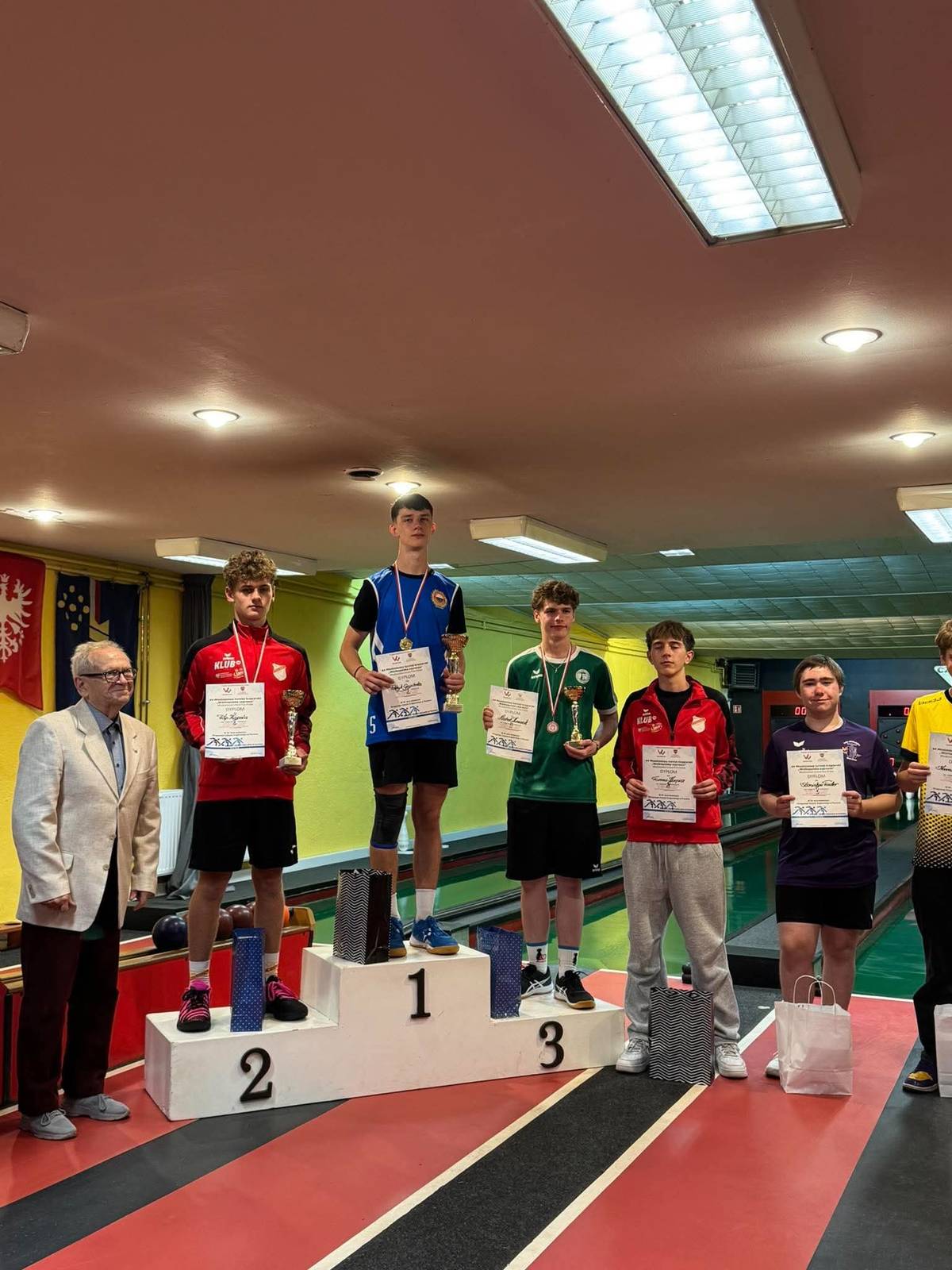 Na zdjęciu kręglarz Pilicy na podium na pierwszym miejscu zawodów Młodzieżowego Grand Prix. Udekorowany medalem, w rękach trzyma dyplom. Podium ustawione na torze kręgielni