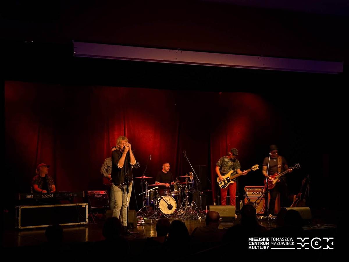 Na zdjęciu Jacek Dewódzki z zespołem Dżem Session podczas koncertu w MCK Tkacz. Przyciemnione, czerwone światło scenicznie, obok wokalisty pełny skład rockowy muzyków