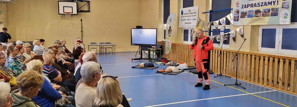 Na zdjęciu ratownik medyczny omawia udzielanie pierwszej pomocy po wypadku drogowym. Wykład odbywa się na sali gimnastycznej, obok ratownika fantomy i tablica edukacyjna. Wykładu słuchaja seniorzy, którzy siedzą na krzesłach na sali gimnastycznej