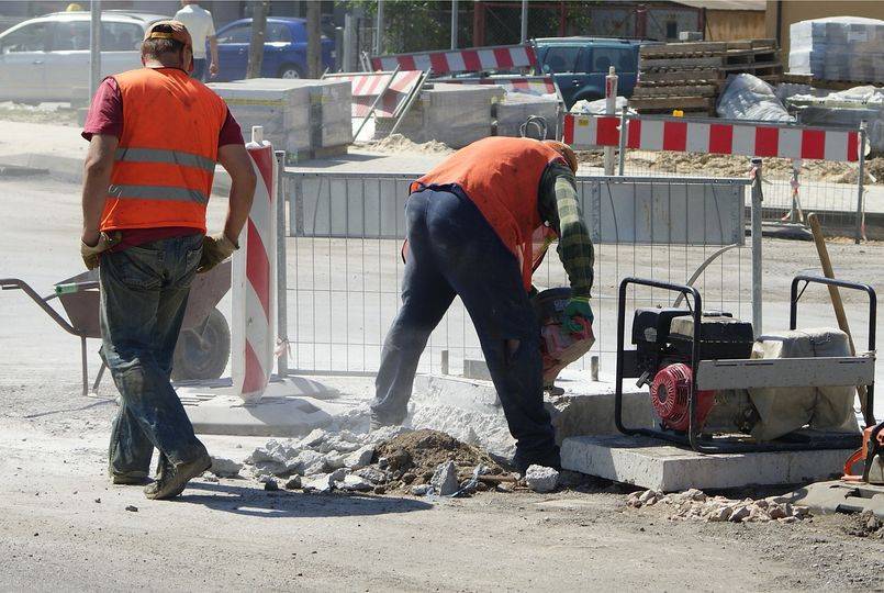 Drogowcy ubrani w pomarańczowe kamizelki wykonują prace drogowe. Na jezdni znajdują się narzędzia m.in. taczka oraz barierki ochronne i palety z kostką. W tle widać samochody.