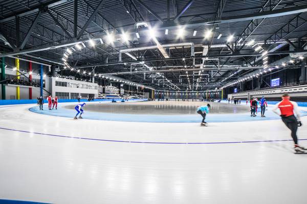 Wnętrze hali lodowej Areny Lodowej z torem łyżwiarskim, po którym jeżdżą łyżwiarze szybcy w kolorowych strojach; w tle widoczne trybuny i oświetlenie sufitowe.