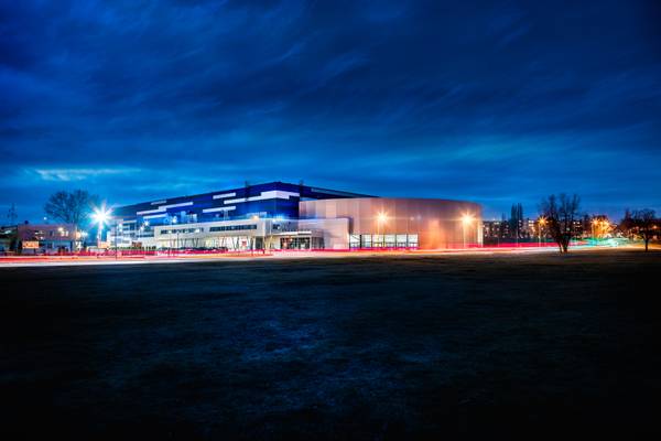 Nocny widok na budynek Areny Lodowej w Tomaszowie Mazowieckim, oświetlony niebieskimi i białymi światłami, z widocznymi smugami świateł przejeżdżających samochodów.