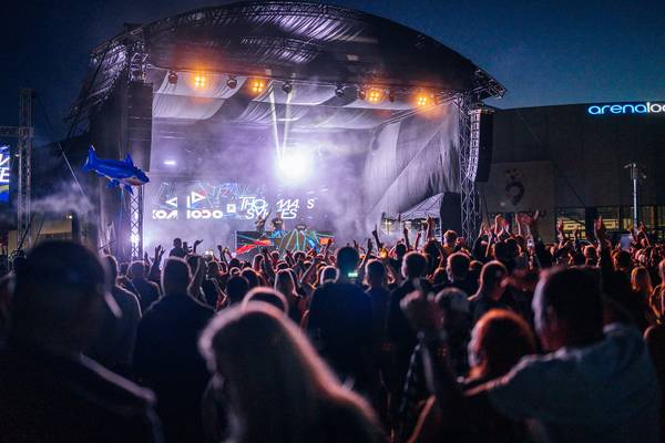 Nocny koncert plenerowy z tłumem bawiących się ludzi, sceną otoczoną dymem i światłami, dekoracyjnym balonem w kształcie rekina oraz napisem „arenalodowa w tle.