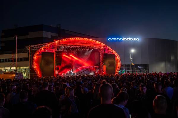 Koncert plenerowy nocą – scena podświetlona na czerwono z występującymi muzykami, tłum widzów z uniesionymi rękami oraz neonowy napis „arenalodowa na budynku w tle.