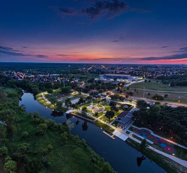 Zdjęcie z drona o zmierzchu przedstawiające oświetlony park nad rzeką z boiskami sportowymi, amfiteatrem, placem zabaw oraz Areną Lodową w tle, na tle różowo-niebieskiego nieba.