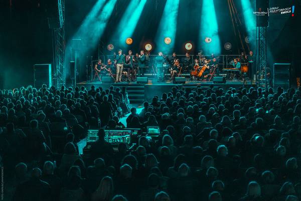 Koncert orkiestry symfonicznej z wokalistami na scenie oświetlonej na turkusowo, przed liczną publicznością wypełniającą widownię.