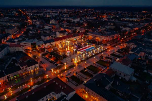Zdjęcie z drona przedstawiające oświetlony jarmark bożonarodzeniowy na placu miejskim nocą, z choinką, karuzelą i świątecznymi iluminacjami w kształcie reniferów i aniołów.