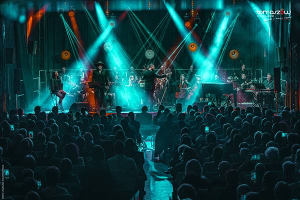 Koncert noworoczny orkiestry z solistą w kapeluszu i dyrygentem na scenie oświetlonej na turkusowo, przed liczną publicznością w sali koncertowej.