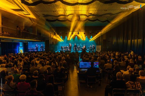 Koncert noworoczny orkiestry symfonicznej z wokalistą na scenie oświetlonej złotym światłem; pełna widownia oglądająca występ, częściowo także na telebimie.