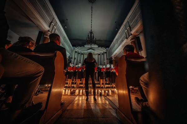 Śpiewaczki chóru trzymające czerwone śpiewniki, oświetlone ciepłym światłem świec podczas koncertu w zabytkowym wnętrzu.