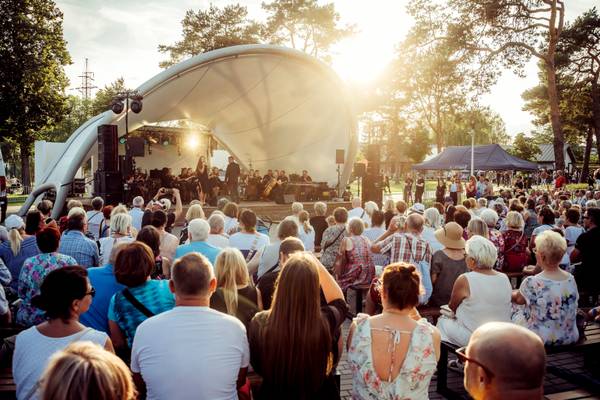 Koncert plenerowy pod białym zadaszeniem sceny w kształcie muszli, z zespołem muzycznym i wokalistką, obserwowany przez tłum siedzącej publiczności o zachodzie słońca.