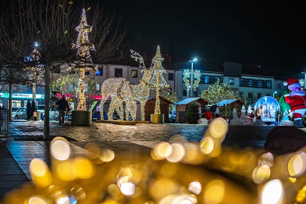 Świąteczna iluminacja na placu miejskim nocą – podświetlone choinki, figura renifera oraz dekoracje Mikołaja, w tle kamienice i domki jarmarczne.