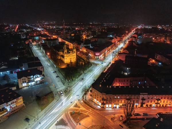 Nocny widok z drona na oświetlone ledowo skrzyżowanie ulic w centrum miasta z zabytkowym kościołem oraz okolicznymi kamienicami.
