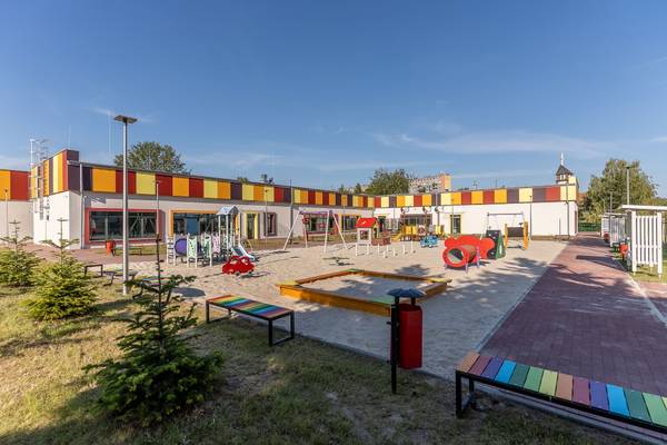 Kolorowy plac zabaw przy żłobku z piaskownicą, zjeżdżalniami i huśtawkami, na tle budynku w żółto-czerwone pasy.
