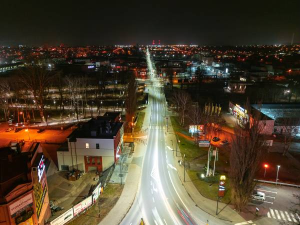 Nocny widok z drona na prostą, oświetloną ulicę miejską ze smugami świateł samochodów, otoczoną zabudową i drzewami.