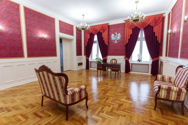 Wnętrze sali ślubów Urzędu Stanu Cywilnego z czerwonymi zdobionymi ścianami, godłem Polski, żyrandolami oraz zabytkowymi fotelami i biurkiem.