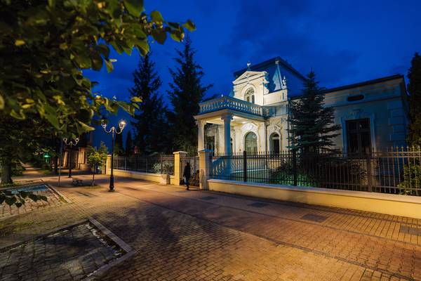 Oświetlony nocą zabytkowy pałacyk z balkonem i kolumnadą, ogrodzony metalowym płotem, przy alei z latarniami.