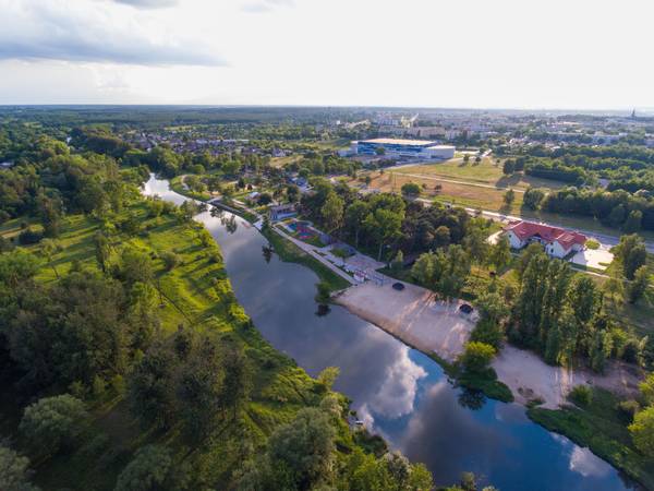 Zdjęcie z drona przedstawiające zakole rzeki z piaszczystą plażą, przystanią i terenami zielonymi, w tle zabudowa miejska oraz hala Areny Lodowej.