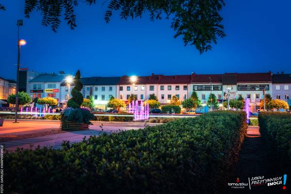 Wieczorny widok na plac miejski z kolorowo podświetloną fontanną (różowo-niebieską) oraz rzędem kamienic ze sklepami w tle.