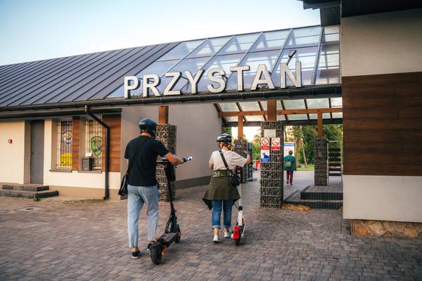 Dwie osoby na hulajnogach elektrycznych wchodzące pod zadaszenie z napisem „PRZYSTAŃ prowadzące do terenu rekreacyjnego nad wodą.