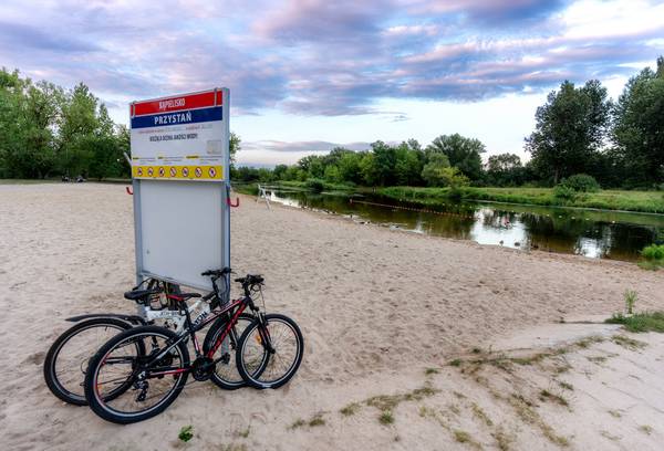 Tablica informacyjna kąpieliska „Przystań na piaszczystej plaży nad rzeką, z opartymi o nią rowerami oraz wytyczonym boją kąpieliskiem w tle.