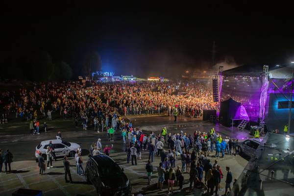 Nocny koncert plenerowy z tłumem licznie zgromadzonej publiczności przed sceną oświetloną fioletowymi światłami, w tle stoiska festiwalowe i samochody.