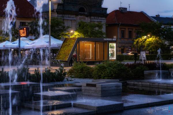 Wieczorny widok na plac miejski z tryskającą fontanną, nowoczesnym pawilonem informacyjnym z zielonym dachem oraz zabytkowymi kamienicami w tle.