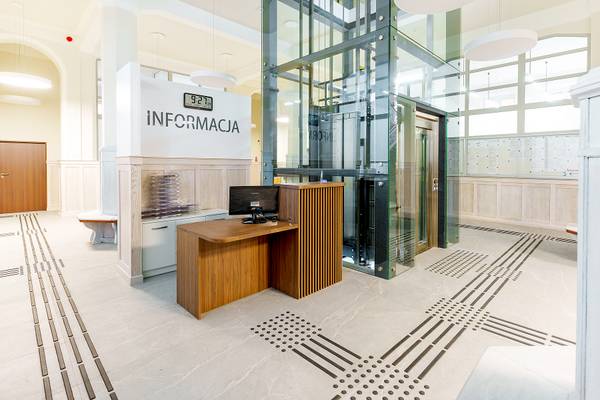 Punkt informacyjny w holu urzędu miasta z napisem „Informacja, szklaną windą w tle oraz posadzką z fakturowymi ścieżkami dla osób niewidomych.