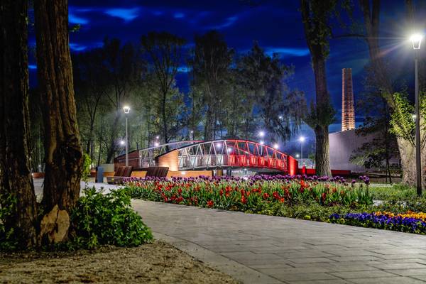 Nocny widok na czerwony kładkowy most nad rzeką w parku Bulwary, oświetlony latarniami, z rabatą kwitnących tulipanów w pierwszym planie oraz kominem fabrycznym w tle.