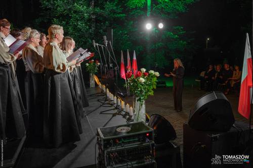 Koncert Chóru Artis Gaudium przed Pomnikiem. Wykonawcy w otoczeniu świateł i biało-czerwonych flag Polski. Przed nimi dyrygent i publiczność. Koncert odbywa się po zmroku, w otoczeniu zieleni.