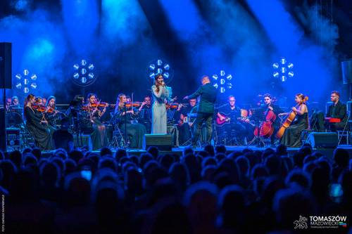 Koncert Katarzyny Zawady z TM Orchestra. Na środku sceny stoi wokalistka, ubrana w długą, białą suknię i śpiewa do mikrofonu. Otacza ją orkiestra. Muzycy siedzą i grają m.in. na skrzypcach i wiolonczeli. Dyrygent stoi tyłem do publiczności