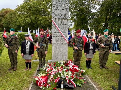 W centrum Pomnik „W Hołdzie Zesłańcom Sybiru”. Przy pomniku warta honorowa harcerzy i wojska. Obelisk zdobi biało-czerwona wstęga i złożone prze delegacje kwiaty. W tle uczestnicy z flagami narodowymi na drzewcach.