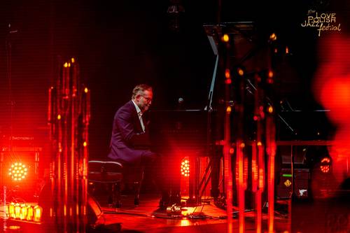 Mężczyzna w garniturze gra na fortepianie podczas Love Polish Jazz Festival. Muzyk siedzi profilem do widza i jest pochylony nad instrumentem. Scena jest oświetlona ciepłym, pomarańczowo-czerwonym światłem.