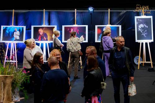 Grupa ludzi zgromadzona w zaciemnionej części Areny Lodowej. W tle na sztalugach stoją fotografie (autorstwa Dariusza Kwapisiewicza) z artystami z  poprzednich edycji festiwalu. Uczestnicy wydarzenia oglądają wystawę i rozmawiają.