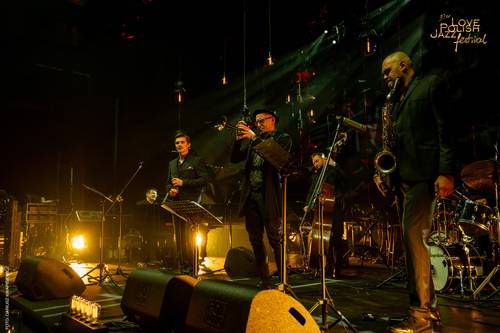 Zespół muzyczny na zaciemnionej scenie gra koncert w czasie festiwalu jezzowego. W centralnej części artyści z instrumentami m.in. saksofonista. Na podłodze stoją elementy nagłośnienia i reflektory - emitujące klimatyczne żółto-pomarańczowe światło.
