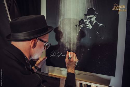 Mężczyzna w okularachi kapeluszu trzyma w dłoni czarny marker, którym podpisuje swój duży, czarno-biały portret. Zdjęcie przedstawia trębacza i pochodzi z poprzedniej edycji festiwalu.