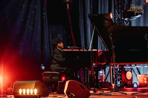 Kobieta gra na fortepianie podczas Love Polish Jazz Festival. Artystka siedzi profilem do widza i śpiewa do mikrofonu. Scena jest oświetlona ciepłym, pomarańczowo-czerwonym światłem. W dolnej części widoczny jest zarys publiczności festiwalowej.