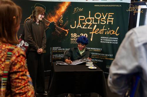 Prz stole nakrytym czarnym materiałem siedzi kobieta, która podpisuje płytę dla stojącej w pobliżu fanki. Za artystką stoi ścianka promocyjna Love Polish Jazz Festival. Na czarnym tle widoczne są złote i białe napisy oraz zarys trąbki.