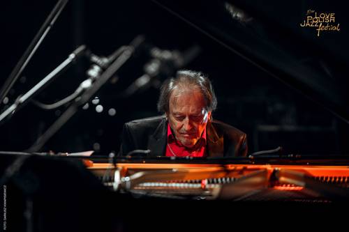 Pianista jezzowy siedzi przy otwartym czarnym fortepianie. Ubrany jest w ciemną marynarkę i czerwoną koszulę. Na sylwetce muzyka jest skoncentrowane nastrojowe oświetlenie.