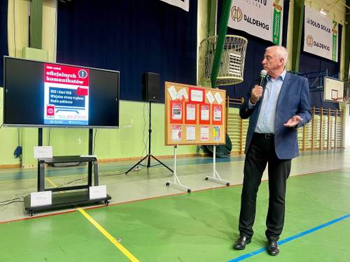 Zastępca Dyrektora Wydziału Kryzysowego i Monitoringu prowadzi wykład na temat rodzajów alarmów podczas Krajowego Treningu Rodzinnego w Szkole Podstawowej nr 1 w Tomaszowiem Maszowieckim. W tle elektroniczna tablica edukacyjna z wyświetlonym postepowanie