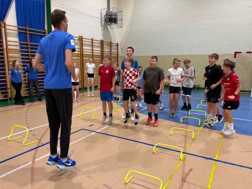 Trening ogólnorozwojowy z Olimpijczykiem Mateuszem Rzeźniczakiem. Uczniowie ćwiczą w strojach gimnastycznych i słuchają wskazówek trenera