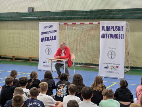 Spotkanie z Olimpijczykiem Marianem Woroninem na sali gimnastycznej. Olimpijczyk prezentuje medal olimpijski i opowiada o swojej karierze sportowej. Spotkanie odbywa się na sali gimnastycznej Szkoły Podstawowej nr 6